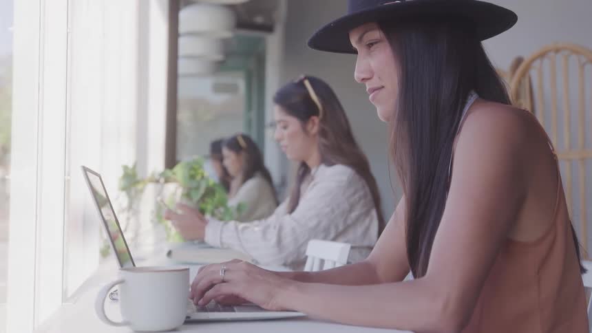 A woman types on a laptop by the window in a bright caf while others sit nearby