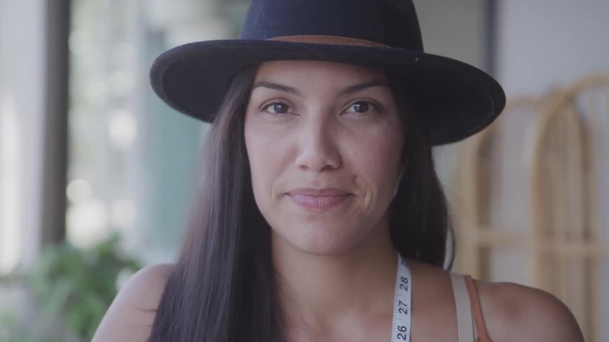 A woman in a widebrimmed hat smiles softly at the camera in a welllit indoor setting