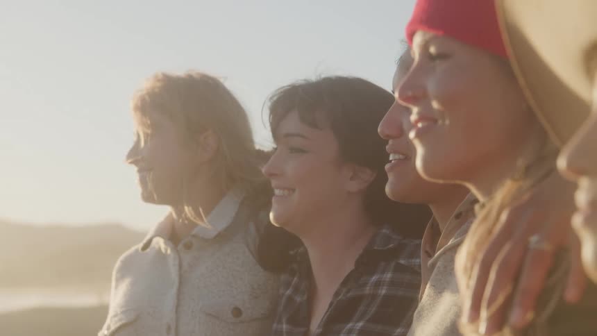 A group of female friends stands together in profile smiling as they look out at a view bathed in warm golden sunlight