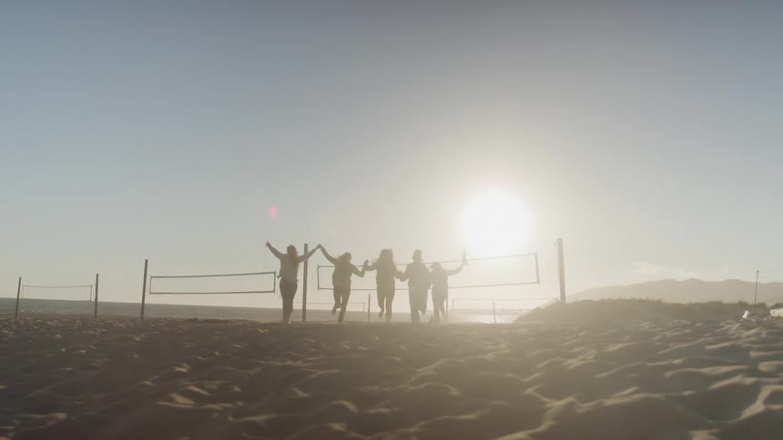 Five friends run barefoot on the beach holding hands and kicking up sand in the sunset light