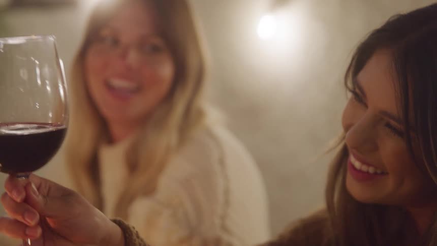 Two women smile and laugh together while holding a glass of red wine in a warmly lit setting
