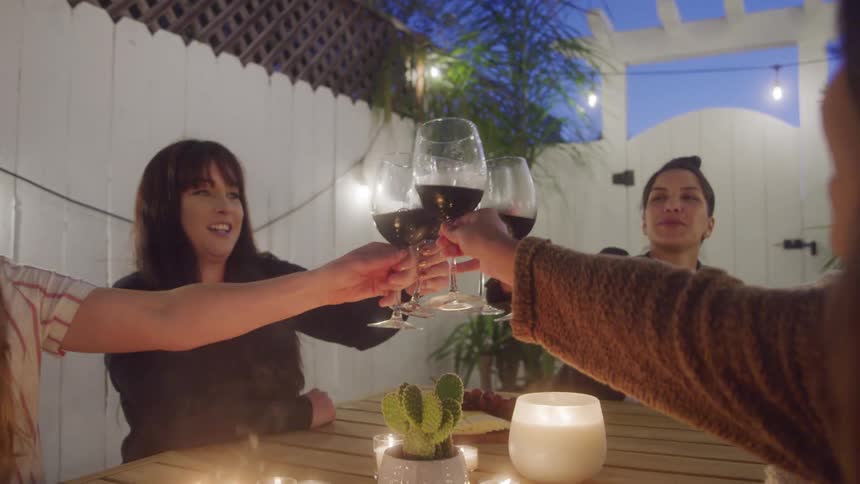 Female friends gathered around patio table by candlelight toasting with wine glasses