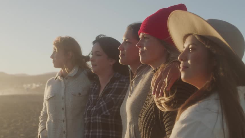 The woman have their arms across each others shoulders on the beach