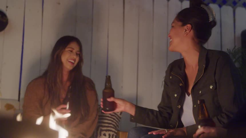 Two women smile and toast with beers while sitting by a fire in an outdoor setting at night