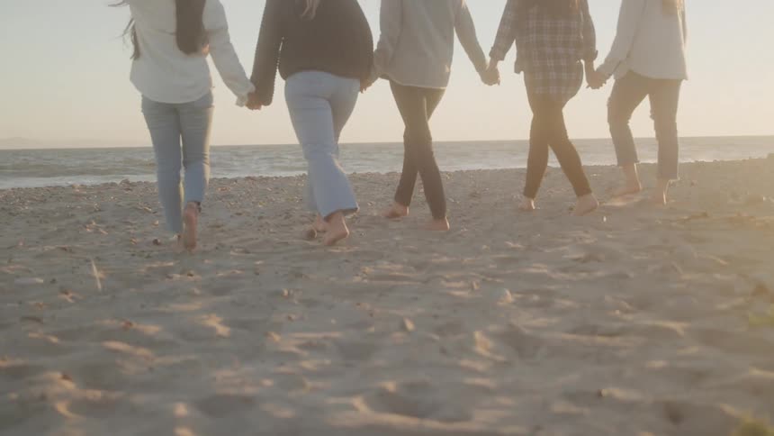 A group of five friends holding hands walk barefoot on the sand toward the ocean at sunset