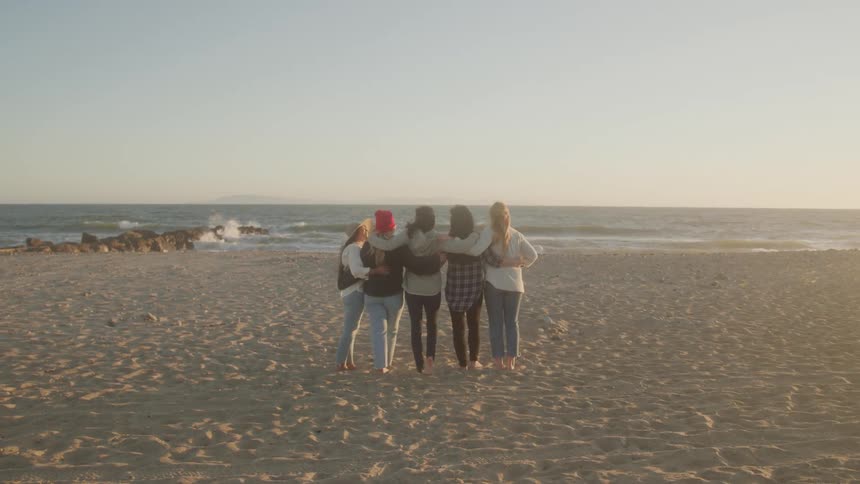 Five people stand side by side on a sandy beach facing the ocean at sunset