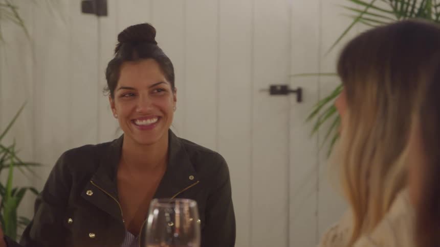 Three woman sit at a table smiling and talking over drinks in a warmly lit setting