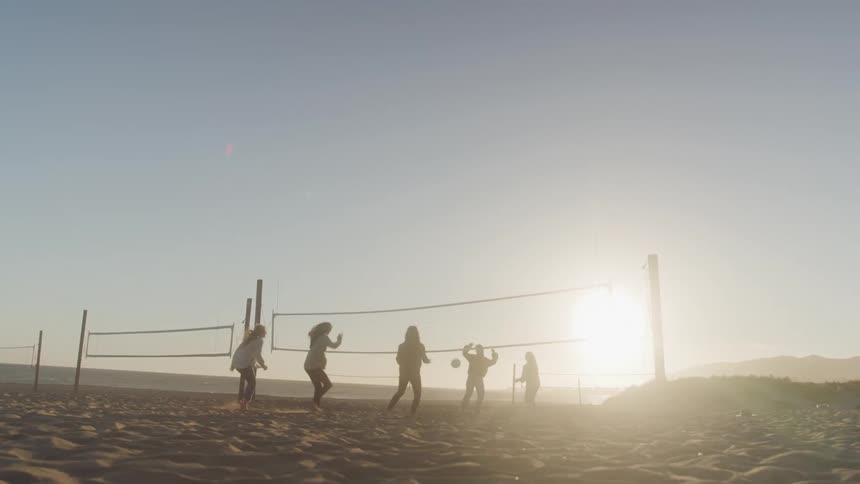 A group of friends play volleyball on a sandy beach at sunset with the sun low on the horizon