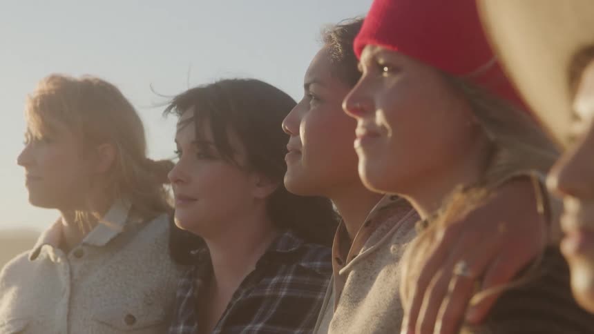 Four young women stand outdoors under the sun holding each other by the shoulders as the wind blows through their hair