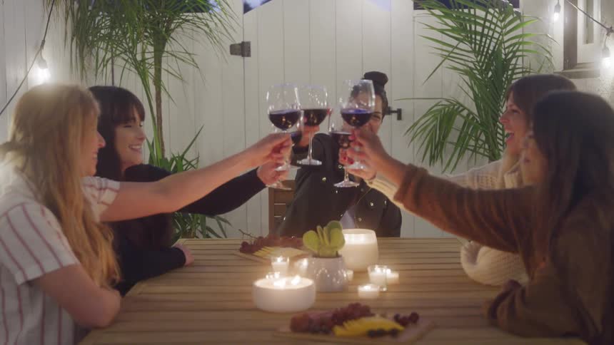Friends clink red wine glasses around a candlelit wooden table topped with a charcuterie board beneath string lights and potted palms