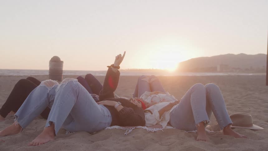 A group of friends lies on the beach at sunset pointing at the sky and enjoying the moment