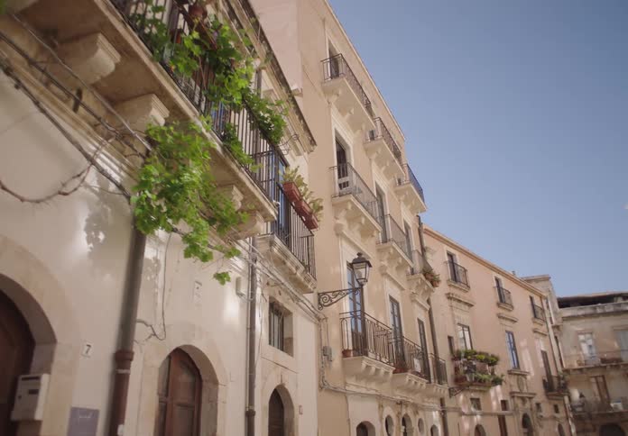 A row of charming historic buildings with balconies lines a narrow street under a clear blue sky