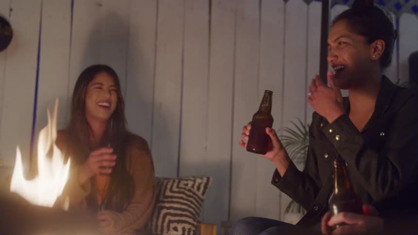 Two women sit outdoors at night laughing and enjoying drinks together