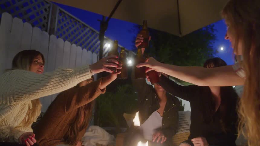 A group of women raise their drinks in a cheerful toast around a fire pit at night