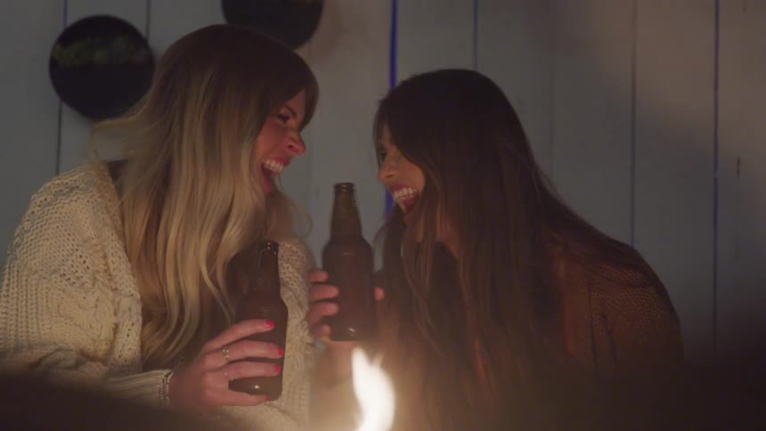 Two women happily clink beer bottles while laughing together