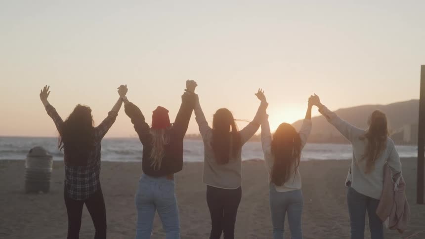 Five women stand on a beach holding hands in the air facing the ocean at sunset
