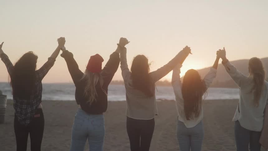 Back of five women holding hands and throwing hands in the air while standing by the beach at sunset