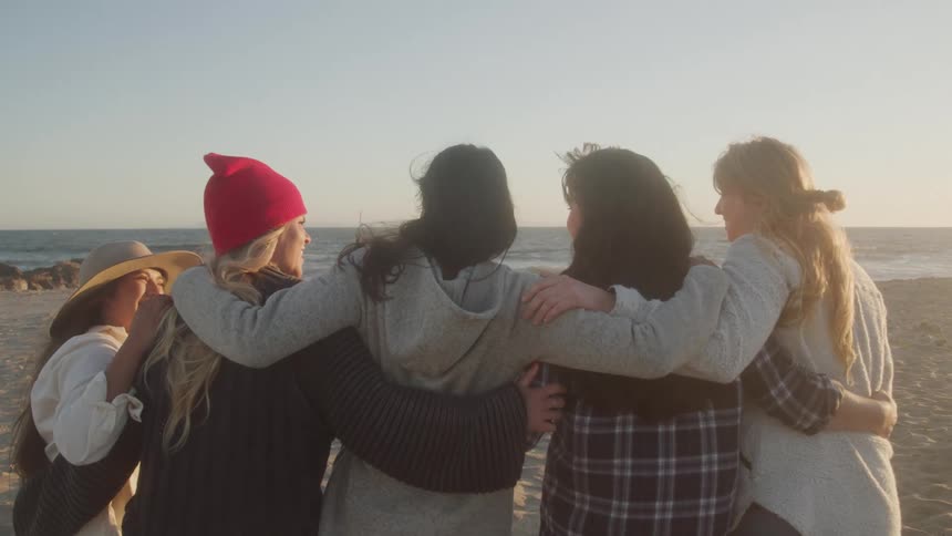 Five women stand arminarm on the beach facing the ocean at sunset