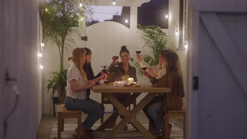 Group of women toast with wine glasses at a cozy candlelit outdoor dinner table under string lights