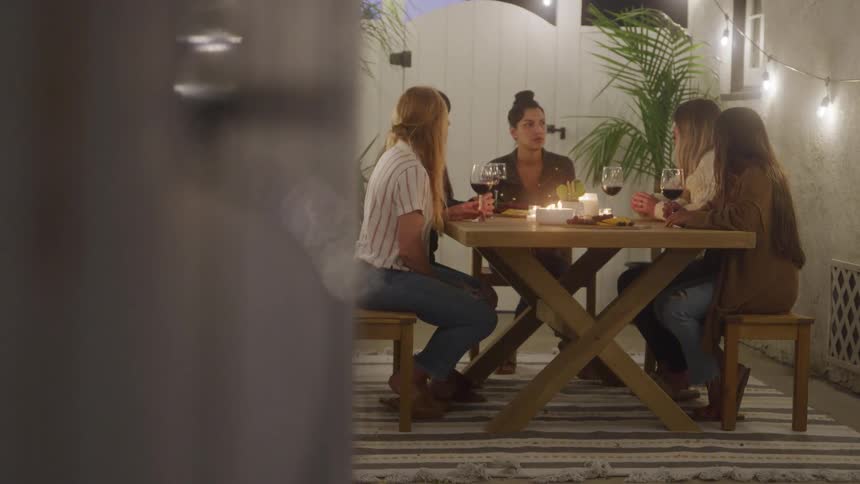Five women sit at a warmly lit outdoor table engaged in conversation over wine