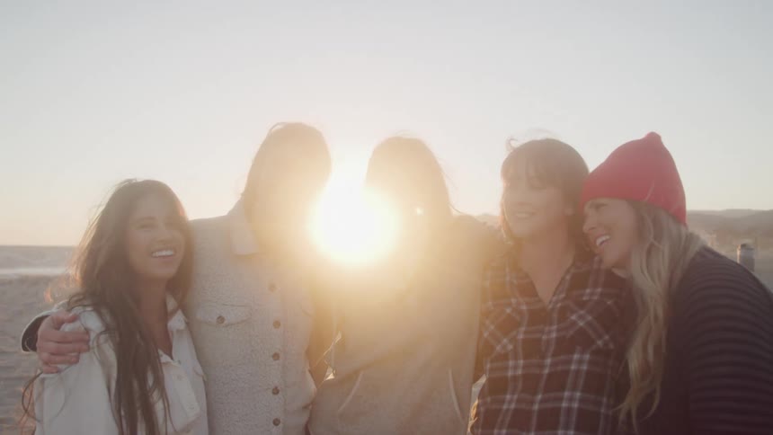 Five female friends stand side by side on the beach smiling on a sunny day