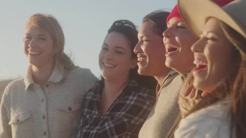 A group of five women laughing while standing side by side on a sunny day