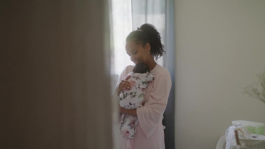 A woman gently holds a swaddled baby close to her chest in a softly lit nursery