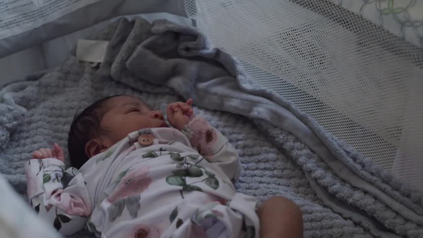 A baby lies peacefully in a crib while gently holding an adults finger