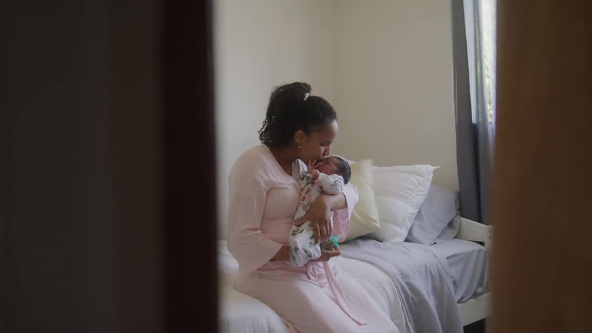 A mother lovingly kisses her swaddled baby while sitting on a bed in a softly lit room