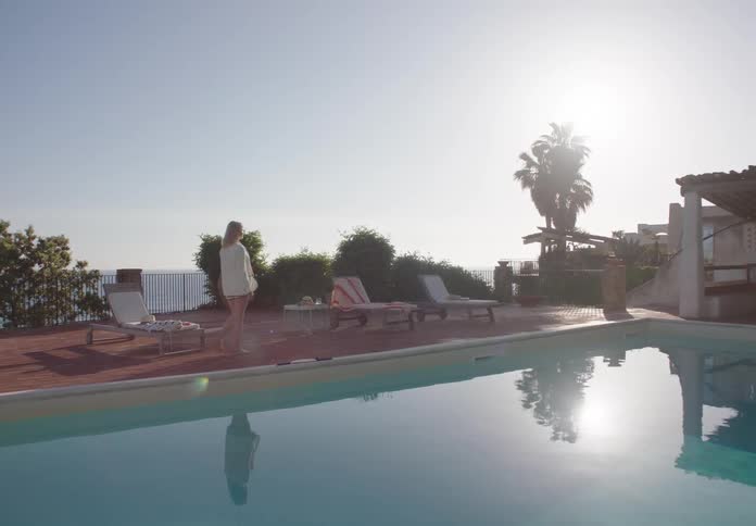 A woman walks by a serene poolside terrace under bright sunlight at a coastal resort