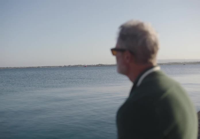 A man with gray hair and sunglasses looks out over a calm body of water