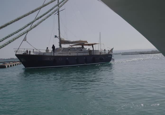 A wide shot of a yacht with people onboard sailing near a dock in calm blue waters