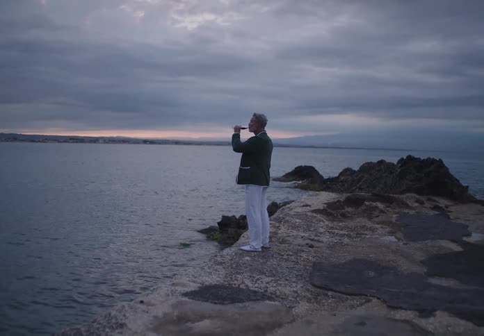An elderly man stands on a rocky shoreline taking a photo of the ocean at dusk