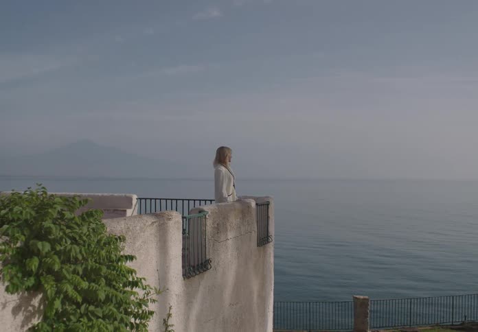 A woman in a light colored jacket stands on a balcony overlooking a wide expanse of ocean and a distant mountain