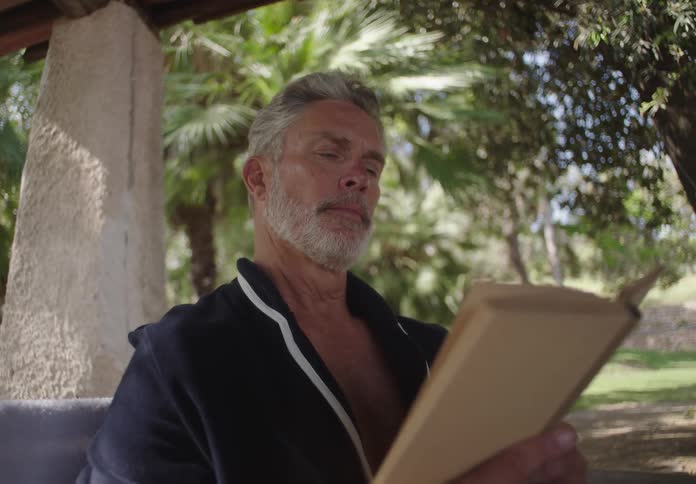 A man with gray hair and a beard wears a dark blue robe and reads a book outdoors