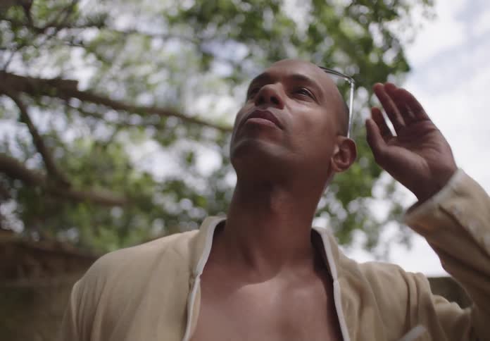 A man with dark skin wearing sunglasses and a light colored jacket looks upwards with tree branches in the background