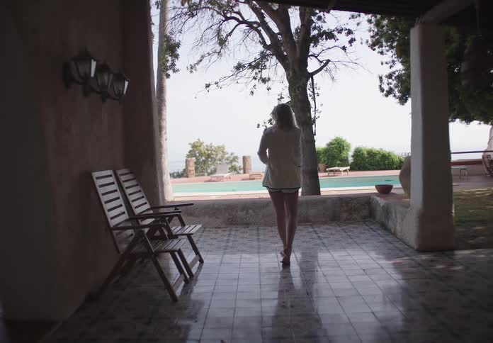 A woman walks alone toward a poolside under a shaded patio