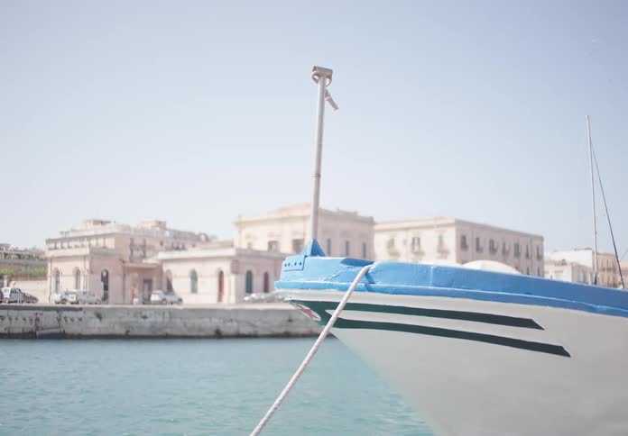 The bow of a small boat rests in the calm water of a harbor with historic buildings lining the shore in the background