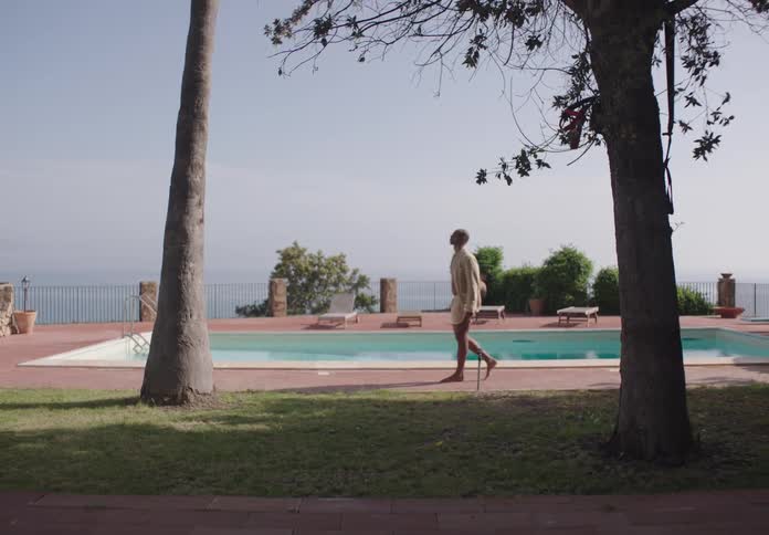 A man walks barefoot near a pool in a sunlit outdoor setting with ocean in the background