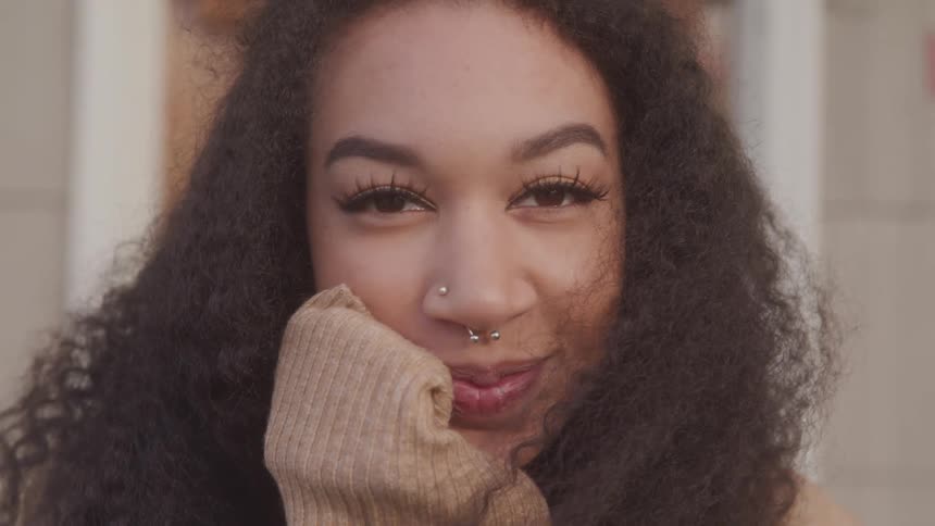 A young woman with a nose piercing rests her hand on her face and smiles