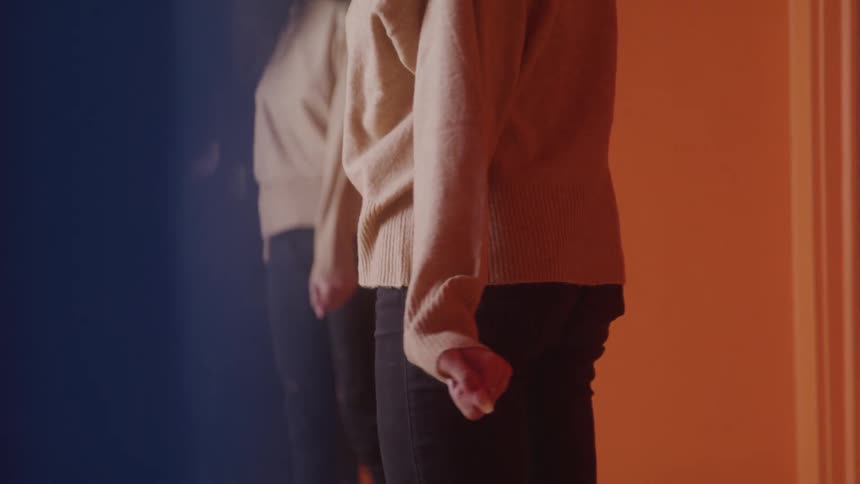 A woman in a light colored sweater and dark pants stands facing a mirror looking at her reflection