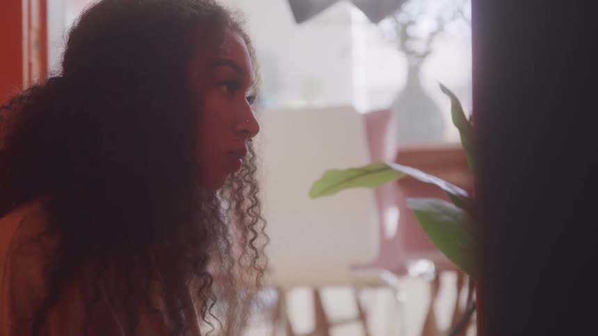 A woman with curly hair looks intently at a plant to her right next to a window