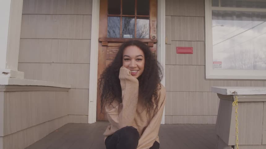 A young woman with curly hair smiles while sitting on the porch of a house with a wooden door