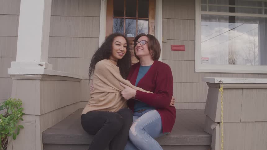A daughter and mother sit side by side on their front porch hugging each other and smiling