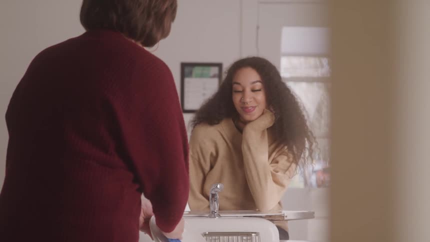 A young woman with curly hair smiles up at another woman in a maroon sweater who is washing dishes in a home kitchen