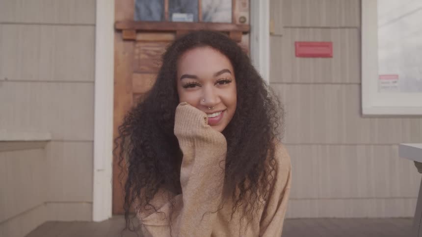 A young woman sits on a porch smiling with her head resting on her hand