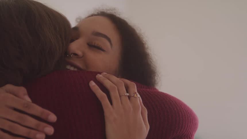 A young woman with curly hair smiles while embracing another woman whose back is to the viewer