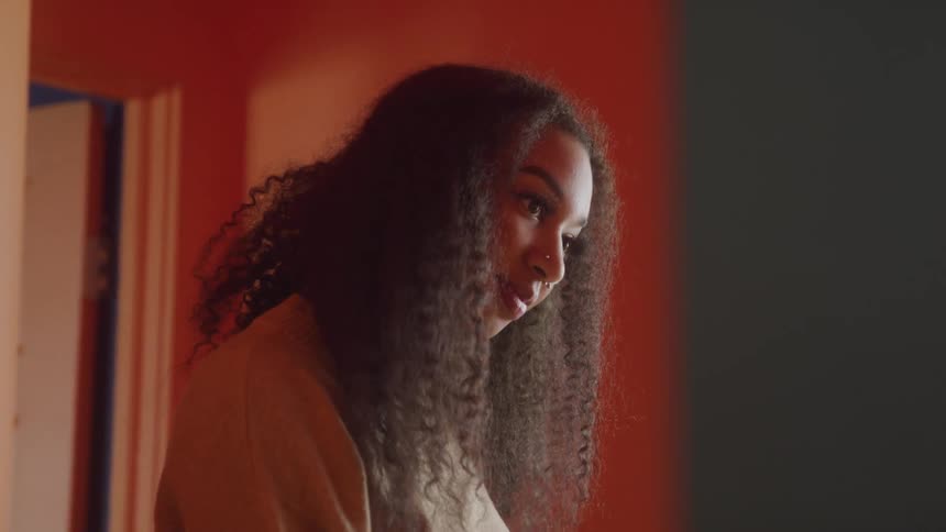 A woman with long curly dark hair looks to her right with a slight smile illuminated by an orange glow from the background