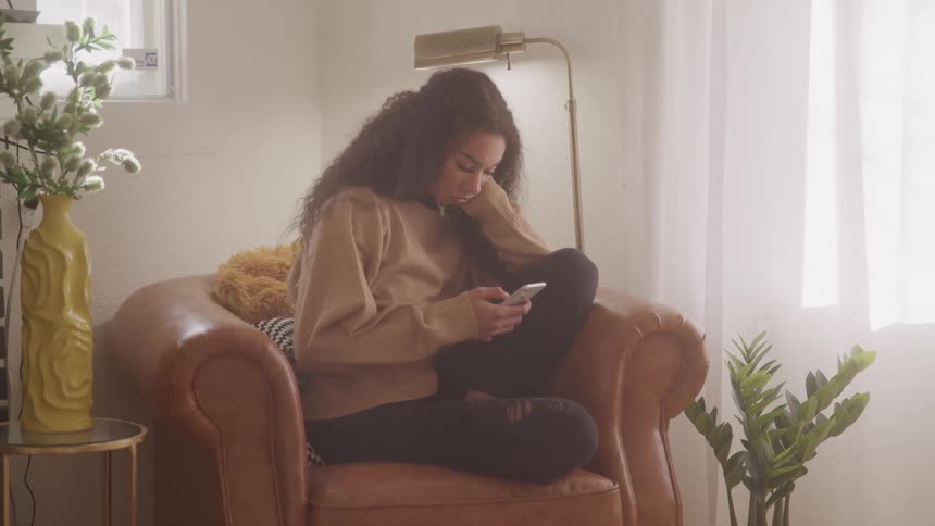 A young woman sits curled up in a cozy armchair focused on her phone in a softly lit room