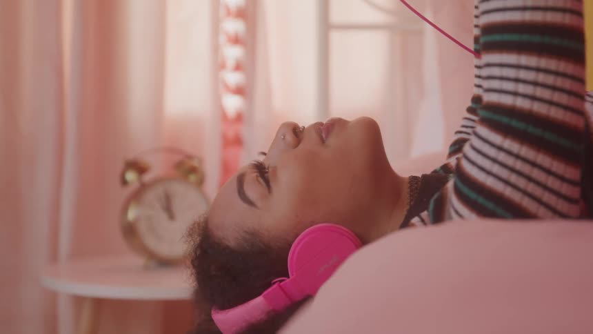 A young woman lies on a bed wearing pink headphones with a smile on her face around midday
